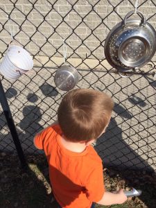 Toddler wearing orange looking at kitchen equipment on a chain fence