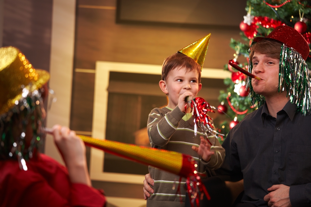 Parents and their child wear party hats and blow into party horns in their living room.